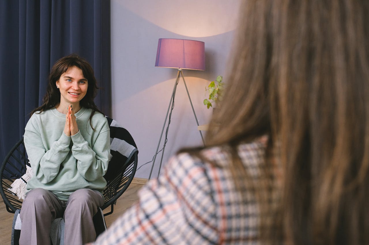 Positive young woman in casual clothes smiling while sitting on chair after psychotherapy session with anonymous female doctor in modern office
